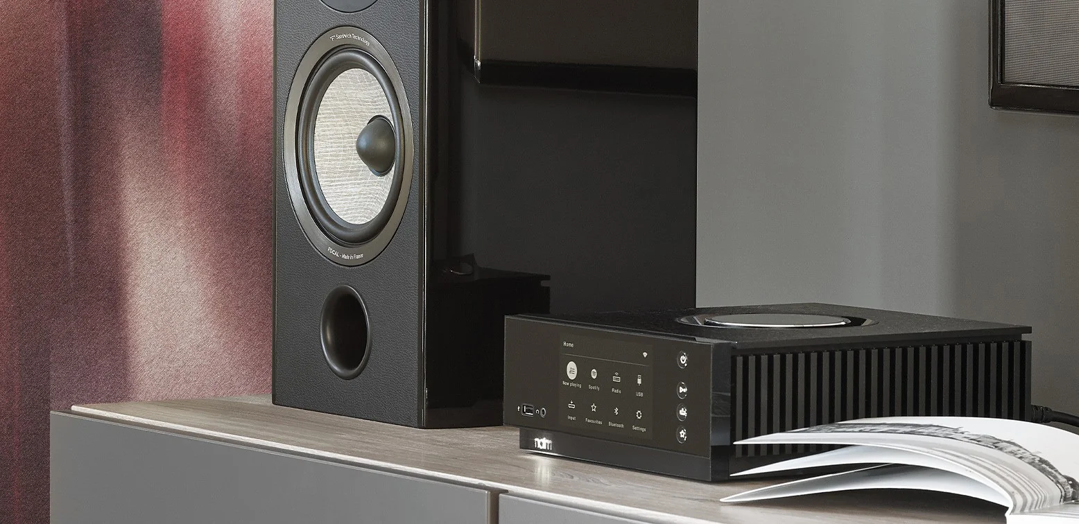 A Naim streaming amplifier on a white sideboard beneath a bookshelf speaker